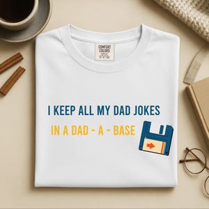 Könnte beinhalten: Ein weißes kurzärmeliges T-Shirt mit einem Comfort Colors-Etikett am Halsausschnitt. Das Shirt zeigt den blauen und gelben Text "I KEEP ALL MY DAD JOKES IN A DAD - A - BASE" sowie eine Grafik einer blauen Diskette mit einem orangefarbenen Pfeilsymbol.