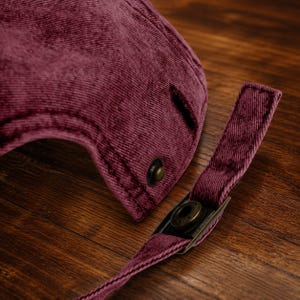 May include: A deep burgundy corduroy baseball cap featuring a textured fabric surface. The back of the cap includes an adjustable strap with a metallic bronze-toned buckle and a matching metallic button detail near the strap attachment point.