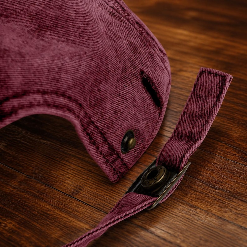 May include: A deep burgundy corduroy baseball cap featuring a textured fabric surface. The back of the cap includes an adjustable strap with a metallic bronze-toned buckle and a matching metallic button detail near the strap attachment point.