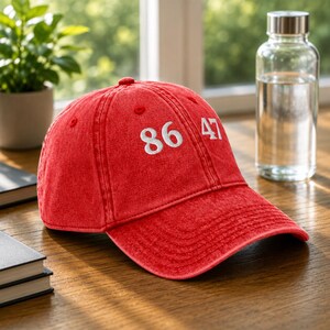 May include: A red baseball cap with a distressed, washed-out texture. The front of the cap features the white embroidered numbers 86 and 47. The cap has a curved brim, a structured crown with ventilation eyelets, and a button on the top center.