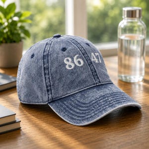 May include: A blue acid-wash denim baseball cap featuring the white embroidered numbers 86 47 on the front panel. The cap has a structured crown, a curved brim with visible stitching, and a button at the top center of the six-panel construction.