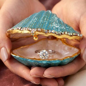 May include: A turquoise seashell-shaped trinket box with a gold-tone rim and embedded iridescent crystals. The hinged lid is open, revealing a silver-tone ring with a large, round, clear faceted stone resting inside the pearlescent interior.