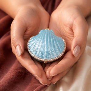 May include: A light blue, shell-shaped trinket box with a metallic gold-toned rim encrusted with small, clear crystals. The surface of the shell features a textured, ribbed design that mimics the natural appearance of a scallop shell.