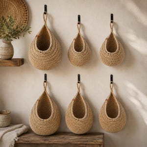 May include: Six teardrop-shaped woven baskets made of natural tan fiber, each featuring a sturdy loop at the top for wall mounting. The baskets are arranged in two rows of three, suspended from black metal hooks against a neutral-colored wall.