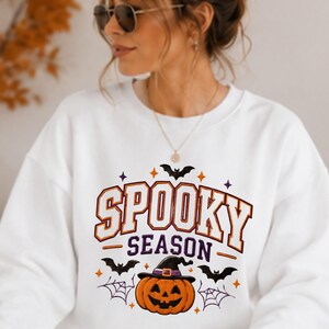 Puede incluir: Una sudadera blanca de cuello redondo con un estampado gráfico que incluye el texto SPOOKY SEASON en letras de bloque naranjas y moradas. El diseño presenta una calabaza central con sombrero de bruja, murciélagos negros y telarañas moradas.
