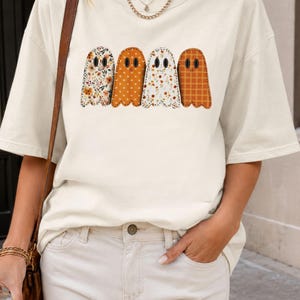 Puede incluir: Una camiseta de manga corta color blanco roto con un estampado gráfico de cuatro fantasmas en fila. Cada fantasma tiene un patrón distinto: floral, de lunares, otro floral y de cuadros, en tonos naranja, marrón y crema.
