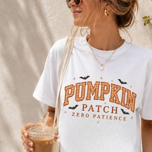Puede incluir: Una camiseta blanca de manga corta con el texto PUMPKIN PATCH en cuadros naranjas y el texto ZERO PATIENCE en marrón. El diseño incluye pequeñas siluetas de murciélagos negros y diminutos detalles de estrellas alrededor de las letras.