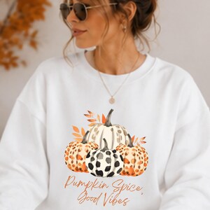 Puede incluir: Una sudadera blanca de cuello redondo con un gráfico de tres calabazas con estampados de leopardo y vaca, acentuadas por hojas naranjas. El texto "Pumpkin Spice, Good Vibes" está impreso en una fuente cursiva naranja bajo la ilustración.