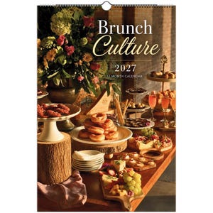 May include: A 2027 wall calendar titled Brunch Culture featuring a photograph of a table spread with doughnuts, biscuits, a charcuterie board with grapes and cheese, glasses of juice, and a large floral arrangement in a vase on a wooden surface.