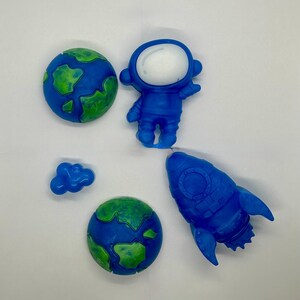 May include: A set of five space-themed items including two spherical globes with blue oceans and green continents, a blue astronaut figure with a white helmet visor, a blue rocket ship, and a small blue cloud-shaped piece on a white background.