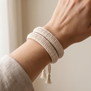 May include: A cream-colored macrame bracelet featuring a double-wrap design with a textured, braided cord pattern. The bracelet is secured with thin, adjustable cord ties that end in small, delicate tassels hanging from the wrist.