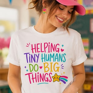 Può includere: Una t-shirt bianca a maniche corte con il testo colorato "HELPING TINY HUMANS DO BIG THINGS" in rosa, viola, verde acqua, verde e giallo. Il design include piccole icone a forma di cuore, stelle e una stella gialla sorridente con scia arcobaleno.