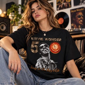 May include: A black short-sleeve t-shirt featuring a graphic of Stevie Wonder wearing sunglasses. The text reads Stevie Wonder, 50, Performances, 50th Anniversary, and Songs In The Key of Life. A circular orange and red design is also visible.