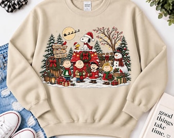 May include: A beige crewneck sweatshirt featuring a graphic print of the Peanuts characters celebrating Christmas. The design includes Snoopy on a doghouse, a snowman, pine trees, gifts, and signs reading Christmas Trees, Hot Cocoa, and Sleigh Rides.