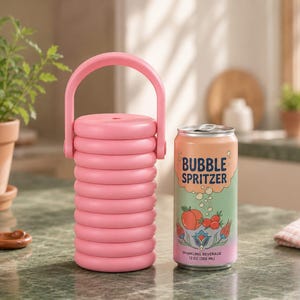 May include: A cylindrical pink beverage cooler with a ribbed texture and a top handle, positioned next to a 12 oz aluminum can labeled Bubble Spritzer with a peach and floral graphic design, featuring a gradient background of green and pink colors.