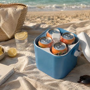 Könnte beinhalten: Eine quadratische, hellblaue tragbare Kühlbox mit weißem Deckel, die vier orangefarbene Fanta-Dosen enthält. Die Kühlbox verfügt über einen zentralen weißen Griffmechanismus und steht auf einer strukturierten Stoffoberfläche neben einer Schale mit Zitronen und einem Buch.