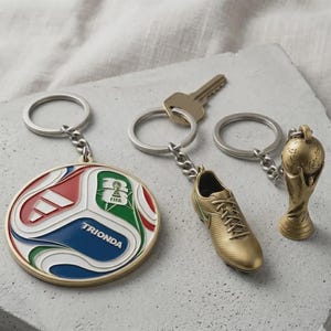 May include: Three metal keychains: a round medallion with red, white, blue, and green enamel featuring the text TRIONDA and a FIFA logo, a gold-toned soccer cleat, and a gold-toned trophy replica, each attached to a silver-colored split ring.