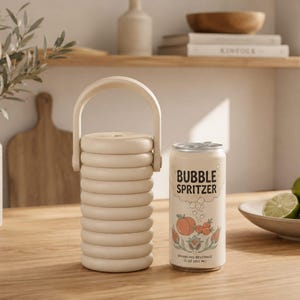 May include: A beige cylindrical beverage cooler with a ribbed texture and a top handle, positioned next to a 12 oz aluminum can labeled Bubble Spritzer with peach and floral graphics, featuring text that reads Sparkling Beverage 12 oz 355 ml.