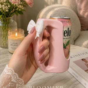 May include: A light pink plastic beverage can holder with a large, integrated handle and a small white fabric bow attached to the side. The holder is designed to fit a standard aluminum can, which is visible inside, featuring a peach oolong label.