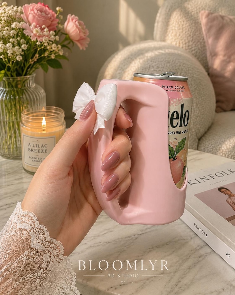 May include: A light pink plastic beverage can holder with a large, integrated handle and a small white fabric bow attached to the side. The holder is designed to fit a standard aluminum can, which is visible inside, featuring a peach oolong label.