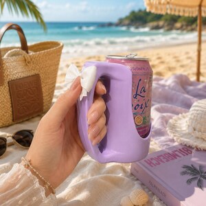 May include: A light purple plastic beverage can holder with a handle and a small white bow attached to the side. The holder contains a pink LaCroix passionfruit sparkling water can. The design features a smooth, curved shape for easy gripping.