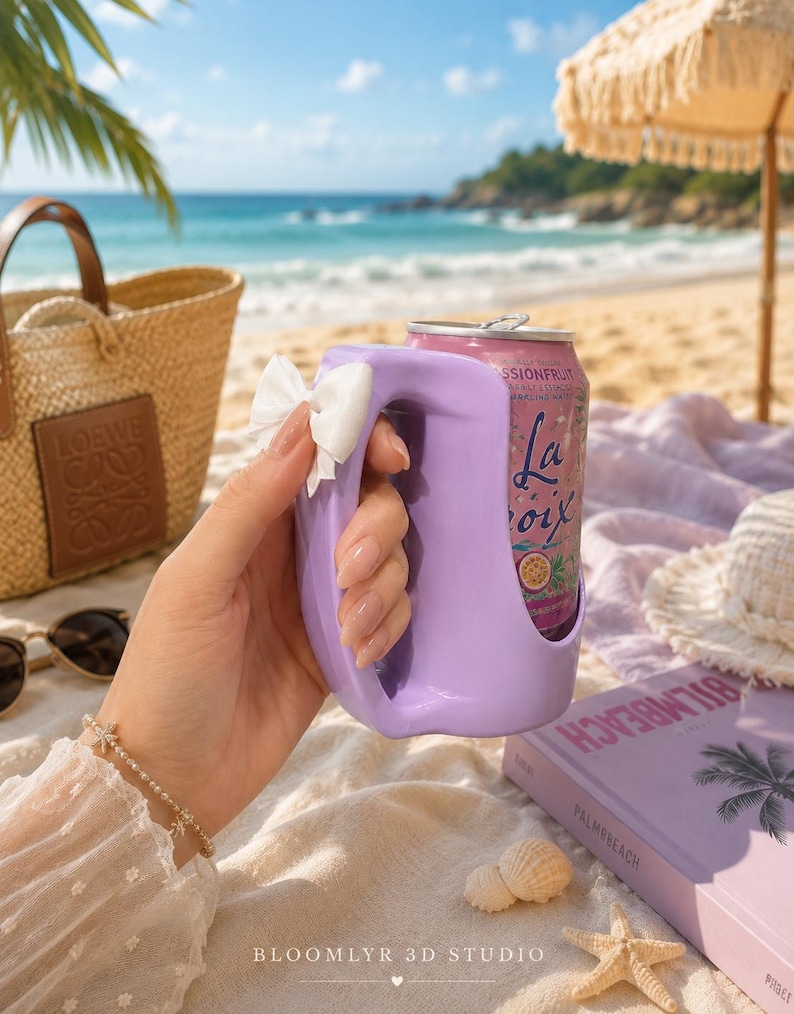 May include: A light purple plastic beverage can holder with a handle and a small white bow attached to the side. The holder contains a pink LaCroix passionfruit sparkling water can. The design features a smooth, curved shape for easy gripping.