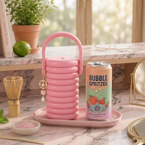May include: A pink cylindrical container with a handle and a small dangling charm, placed on a matching pink oval tray next to a 12 oz can of Bubble Spritzer sparkling beverage. The can features a peach and floral graphic with the text Bubble Spritzer.