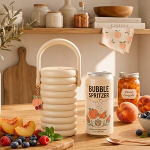 May include: A cream-colored cylindrical container with a handle and a peach-shaped charm, next to a 12 oz can labeled BUBBLE SPRITZER with peach illustrations, a jar of peach compote, fresh peaches, blueberries, raspberries, and a spoon.