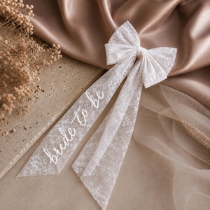 May include: A white lace hair bow featuring two long, trailing ribbons. The left ribbon displays the text "bride to be" in a white cursive script. The bow is constructed from a delicate, semi-sheer floral lace fabric with a soft, intricate pattern.