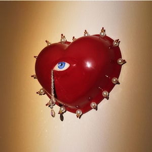 May include: A glossy red heart-shaped object featuring a realistic blue eye embedded in the centre. A single crystal teardrop hangs from the eye. The perimeter of the heart is decorated with small white pearls and gold-toned metal accents.