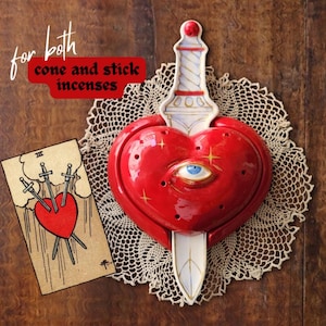 May include: A red ceramic heart incense burner pierced by a white dagger with gold accents and a central blue eye. The text "for both cone and stick incenses" is visible. A tarot card depicting a heart pierced by three swords is placed nearby.