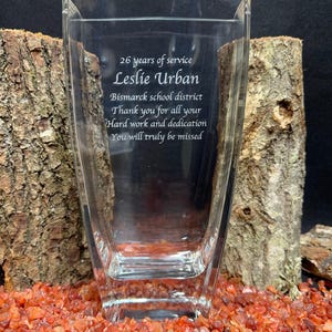 May include: A clear glass vase with an engraved message that reads: 26 years of service, Leslie Urban, Bismarck school district, Thank you for all your hard work and dedication, You will truly be missed. The vase has a rectangular shape.