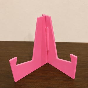 May include: A bright pink display stand constructed from two interlocking flat pieces. The stand features a central vertical support and two angled arms extending outward to hold items, showcasing a smooth, matte finish and a simple geometric design.