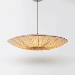 May include: A large, disc-shaped pendant light fixture featuring a beige, fabric-covered shade with fine, radiating pleats. The light is suspended from a thin, vertical metal cord attached to a circular ceiling mount, emitting a soft, warm glow.