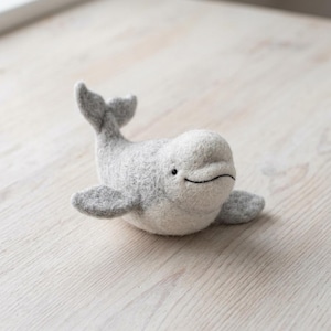 May include: A needle-felted beluga whale figurine crafted from soft, light grey wool fibres. The whale features a rounded head, a small black bead eye, a stitched dark mouth line, two side fins, and a raised tail fin, all in a textured finish.