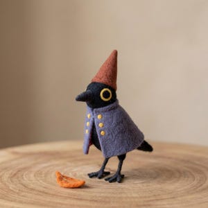 May include: A needle-felted bird figurine with a black body, a pointed rust-colored hat, and a purple cape featuring six small yellow buttons. The bird has large yellow eyes and stands on thin black legs, with a small orange leaf placed nearby.