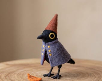 Pode incluir: Uma estatueta de pássaro em feltro com corpo preto, um chapéu pontiagudo cor de ferrugem e uma capa roxa com seis pequenos botões amarelos. O pássaro tem olhos amarelos grandes e apoia-se em patas pretas finas, com uma pequena folha laranja ao lado.