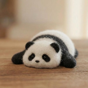May include: A small, fuzzy, needle-felted panda figurine lying flat on its stomach. The panda has a white face and body with black patches around its eyes, black ears, and black limbs. It features a small black nose and two dark, round eyes.