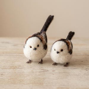 Puede incluir: Dos figuras de aves mito hechas con lana fieltrada. Cada ave presenta un pecho y cara blancos, redondos y esponjosos, con alas de patrones marrones y negros y una cola larga y oscura. Tienen picos negros pequeños, ojos oscuros y patas finas.