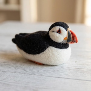Könnte beinhalten: Eine nadelgefilzte Papageientaucher-Figur mit schwarzem Rücken, weißem Bauch und einem markanten Schnabel mit orangefarbenen, gelben und grauen Streifen. Der Vogel hat ein kleines dunkles Auge und einen runden, kompakten Körper aus weicher Wolle.