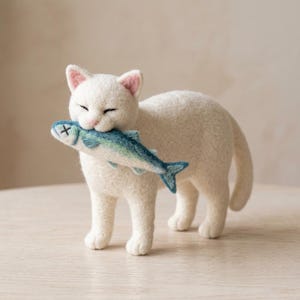Könnte beinhalten: Eine aus Nadelfilz gefertigte weiße Katzenfigur, die auf vier Beinen steht und die Augen geschlossen hat. Die Katze hält einen kleinen blau-grünen Filzfisch im Maul. Der Fisch hat ein mit einem schwarzen Kreuz besticktes Auge und dunkelblaue Flossen.
