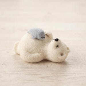 Könnte beinhalten: Eine nadelgefilzte, cremefarbene Eisbärenfigur, die auf dem Rücken liegt, mit einem kleinen, hellgrauen Robbenbaby auf dem Bauch. Der Eisbär hat eine schwarze, gestickte Nase und Augen, während das Robbenbaby winzige schwarze Punktaugen hat.