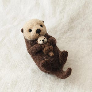 Puede incluir: Una escultura de lana fieltrada de una nutria marina adulta de color marrón oscuro con la cabeza crema, que sostiene a una cría de nutria a juego contra su pecho. Ambas nutrias tienen ojos negros pequeños, narices oscuras y fibras de lana.