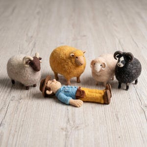 May include: A needle-felted figurine of a person wearing a brown hat, blue shirt, and yellow pants lying down, surrounded by four needle-felted sheep figures: one white with brown face, one mustard yellow, one cream, and one black with horns.