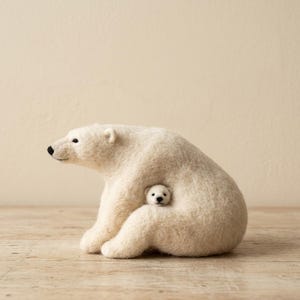 May include: A needle-felted sculpture of a large white polar bear sitting in profile, with a small polar bear cub nestled against its side. Both bears have textured off-white wool bodies, small black bead eyes, and stitched black noses.