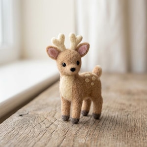May include: A needle-felted deer figurine featuring a light brown body with a cream-colored chest and underbelly. The deer has small antlers, pink-lined ears, black bead eyes, and white spots on its back, standing on four dark brown hooves.