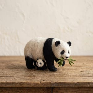 Könnte beinhalten: Eine nadelgefilzte Skulptur eines großen erwachsenen Pandas und eines kleinen Pandababys. Der erwachsene Panda ist weiß mit schwarzen Flecken an Ohren, Augen und Gliedmaßen und hält einen kleinen grünen gefilzten Bambuszweig in seinem Maul.