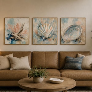 Op de afbeelding: Een set van drie vierkante ingelijste wandkunstwerken met kustthema's. Het linkerpaneel toont koraal en drijfhout, het middelste paneel een grote sint-jakobsschelp en het rechterpaneel een abaloneschelp, in zachte blauwe en beige tinten.