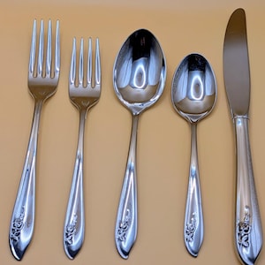 May include: A set of five polished silver-colored flatware pieces arranged on a light orange surface. The set includes two forks, two spoons, and a knife. Each piece features a floral design at the base of the handle. The utensils are reflective, showing the surrounding environment.