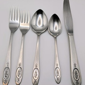 May include: A five-piece stainless steel flatware set consisting of a dinner fork, a salad fork, a dinner spoon, a teaspoon, and a dinner knife. Each handle features an oval-shaped embossed floral design with decorative scrollwork accents.